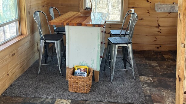 Un interior de cabaña acogedor con una mesa de comedor de madera, sillas metálicas, una cesta pequeña y una ventana con vistas a árboles.