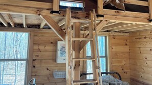 Interior de una cabaña de madera con una cama en el altillo, una escalera, un escritorio y una silla.