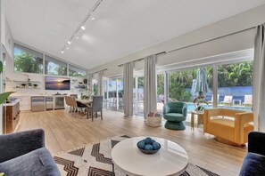 A spacious living area with a dining table, chairs, and a coffee table. There is a kitchen area with a refrigerator and a TV. Large windows provide views of the outdoors. - Beachfront Bliss: 6 Bedroom Home with Heated Pool (Fort Lauderdale)