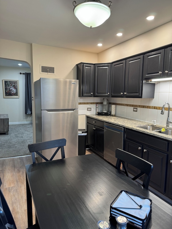 A modern kitchen with dark cabinets, a stainless steel refrigerator, a granite countertop, and a dining area with a wooden table and chairs.