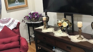 Un salón con un mueble bajo para televisión de madera oscura, un sillón rojo con lunares, un cuadro enmarcado y una mesita con un jarrón de flores y velas.
