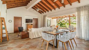 A spacious living area with a wooden ceiling, a dining table with wicker chairs, a sofa, and a TV. - Las Quintas 6B - 2BR Chic Condo on Golf Course/SDC Hotel Zone, Beach & Dining. (San José del Cabo)
