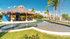 A swimming pool with a thatched-roof structure, palm trees, and a clear blue sky. - Las Quintas 6B - 2BR Chic Condo on Golf Course/SDC Hotel Zone, Beach & Dining. (San José del Cabo)