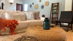 A living room with a white sofa, a round woven ottoman, a teal vase, and a black chair. - Las Quintas 6B - 2BR Chic Condo on Golf Course/SDC Hotel Zone, Beach & Dining. (San José del Cabo)