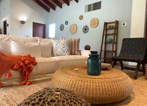 A living room with a white sofa, a round woven ottoman, a teal vase, and a black chair. - Las Quintas 6B - 2BR Chic Condo on Golf Course/SDC Hotel Zone, Beach & Dining. (San José del Cabo)