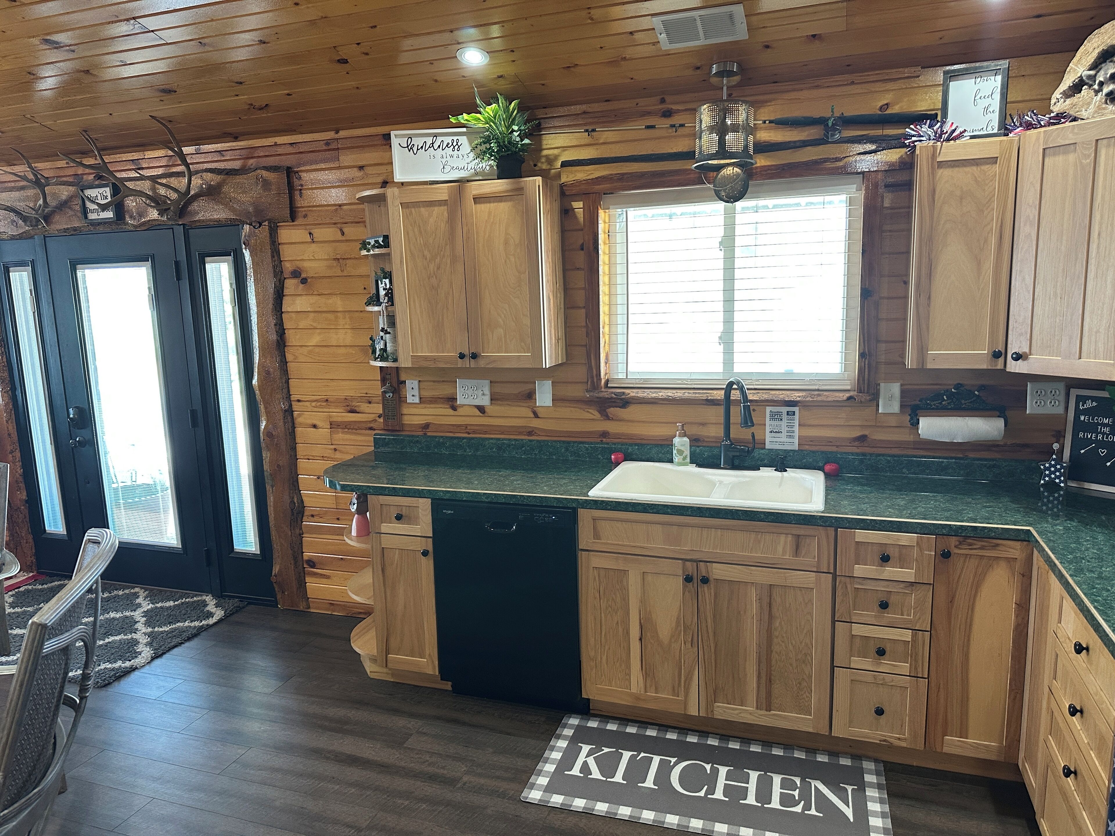 A wooden cabin kitchen with a granite countertop, wooden cabinets, a built-in dishwasher, and a window with blinds.