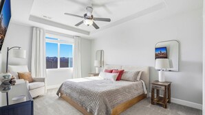 A bedroom with a large bed, a ceiling fan, a nightstand, a lamp, and a view of the city through the window. - Haven in the Desert - Kennewick spacious home (Kennewick)