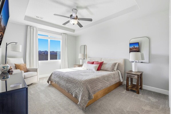 A bedroom with a large bed, a ceiling fan, a nightstand, a lamp, and a view of the city through the window. - Haven in the Desert - Kennewick spacious home (Kennewick)