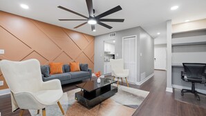 A modern living room with a glass coffee table, white armchairs, a dark ceiling fan, and a kitchen area in the background.