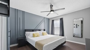 A bedroom with a bed, a ceiling fan, a nightstand, a dresser, a window with curtains, and a wall-mounted mirror. - Premium Townhome #A w/ 2 King Beds & Luxury Baths (Peachtree Corners)