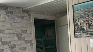A room with a brick wall, a wooden chair, a tiled floor, a door leading to a bathroom, and a framed picture on the wall. - Logement très spacieux.portail un code digital.bte à clé. Animals refused. (Dangé-Saint-Romain)