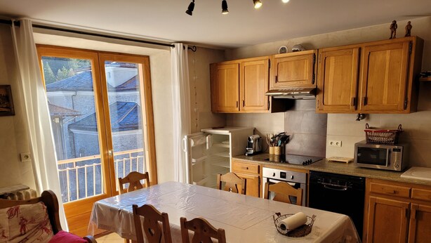 Una cocina con armarios de madera, una mesa de comedor con sillas, un refrigerador, un microondas y una ventana con cortinas.