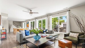 A modern living room with a sofa, coffee table, and ottoman, a dining area with a table and chairs, and large windows with curtains. - Lakefront Oasis w/ Private Dock + Paddle Boards (Lake Ozark)