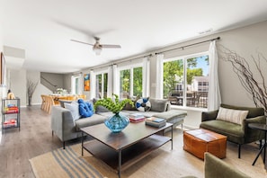 Ein modernes Wohnzimmer mit Sofa, Couchtisch und Hocker, ein Essbereich mit Tisch und Stühlen sowie große Fenster mit Vorhängen.