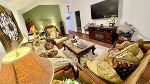 A living room with a sofa, armchairs, a coffee table, and a TV. There is a cross on the wall and a door leading to another room. - Casa de Capucine: Timeless Elegance & Grand Interiors. (Humble)