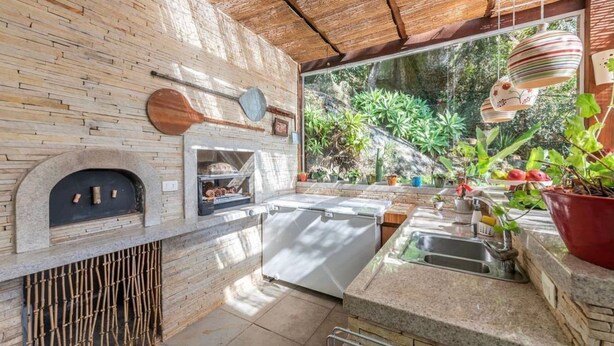 Cozinha externa com parede de pedra, forno a pizza e grande janela com vista para o verde.