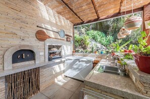 An outdoor kitchen with a stone wall, a pizza oven, and a large window overlooking greenery.