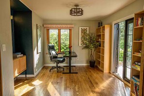 Ein Zimmer mit Holzboden, einem schwarzen Bürostuhl, einem Schreibtisch mit einer Vase und einer Topfpflanze, einem Bücherregal und einem Fenster mit Blick ins Grüne.