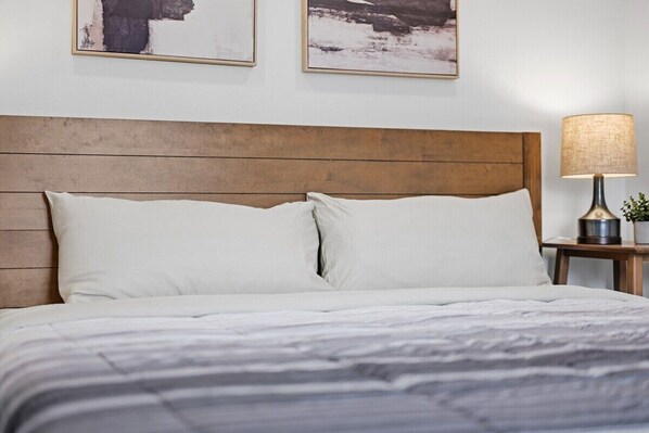 Cama bem arrumada com cabeceira de madeira, almofadas brancas e colcha listrada. Há um abajur e um quadro na parede.