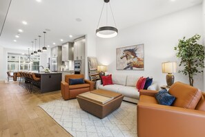 Ein modernes Wohnzimmer mit einer Couch, einem Couchtisch und Sesseln. Im Hintergrund befindet sich eine Küche mit Schränken und einem Essbereich.