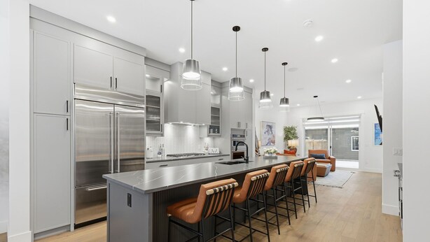 A modern kitchen with stainless steel appliances, a large island with bar stools, pendant lighting, and a view to the living area. - Modern Home with Spa Bath & Garage Altadore (Calgary)