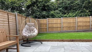 Een terras met een houten bank en een stoel met kussens, een vloer van stenen tegels en een houten hek op de achtergrond.