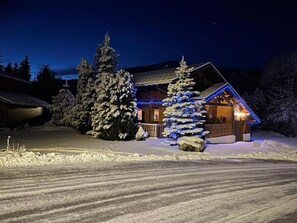 Una cabaña cubierta de nieve con fachada de madera, rodeada de árboles cargados de nieve e iluminada por luces azules.