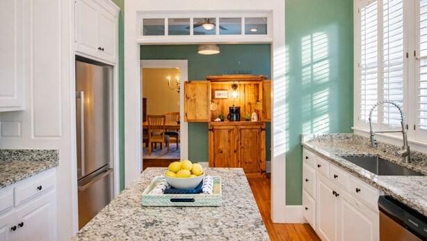 A kitchen with white cabinetry, a granite countertop, a stainless steel refrigerator, and a wooden dining area visible through an archway.
