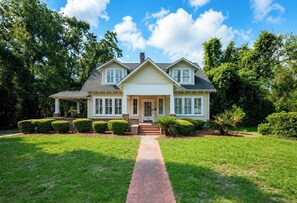 A two-story house with a front porch, a brick pathway leading to the entrance, and well-maintained front yard with trimmed bushes. - Steps to VSU • Game Day HQ • Historic Home • Private Pool • Sleeps 10 (Valdosta)