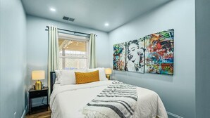 Une chambre à coucher avec un lit, deux tables de chevet avec des lampes, une fenêtre avec des rideaux et un mur orné d’une œuvre d’art colorée.