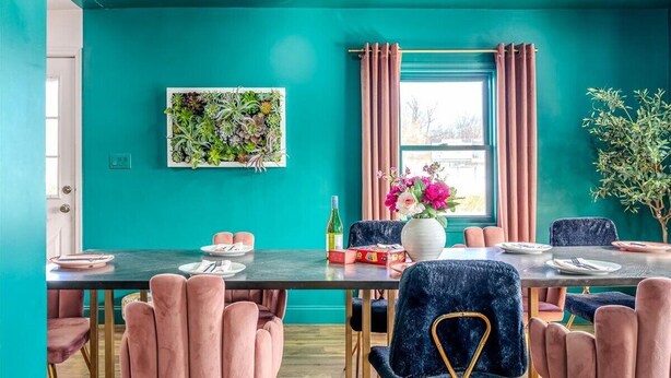 Une salle à manger aux murs de couleur turquoise, avec des chaises roses, une table dressée pour quatre personnes et une fenêtre ornée de rideaux roses.