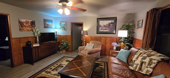 A cozy living room with a television, wooden furniture, a ceiling fan, and various wall art.