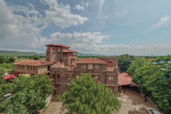 A large, multi-story stone building with a red-tiled roof, surrounded by lush greenery and a hilly landscape in the background. - Seraibagh Ranthambore (Sawai Madhopur)