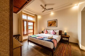 A hotel room with a bed, a ceiling fan, a window with curtains, a bedside table with a lamp, and a rug. - Seraibagh Ranthambore (Sawai Madhopur)
