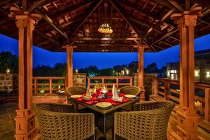 A covered outdoor dining area with a table set for four, surrounded by wicker chairs, under a wooden pergola with a hanging light fixture. - Seraibagh Ranthambore (Sawai Madhopur)
