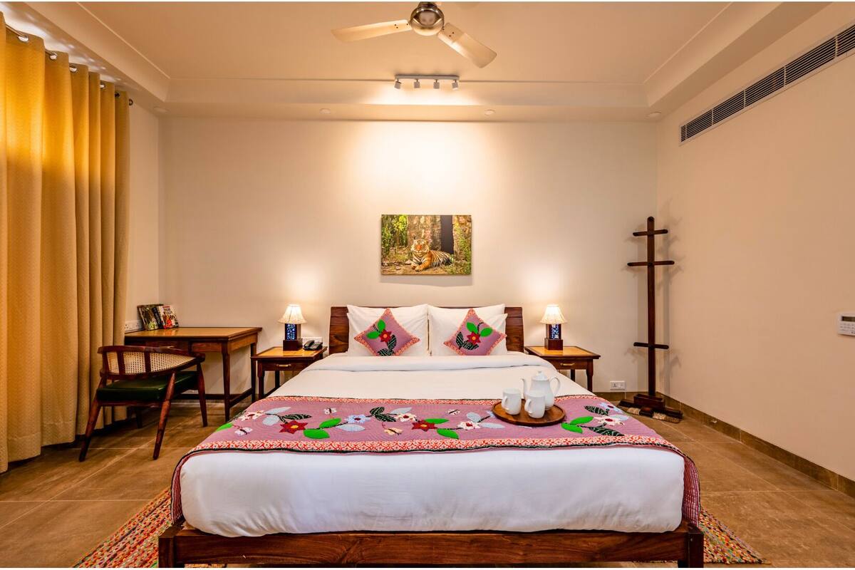 A hotel room with a bed, two bedside tables, a desk, a chair, a ceiling fan, a painting, and a cross on the wall.