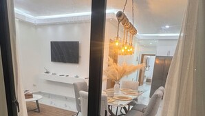 Ein modernes Wohnzimmer mit Glastür, die sich öffnet, einem Essbereich mit weißem Tisch und grauen Stühlen sowie einer ungewöhnlichen Hängeleuchte.