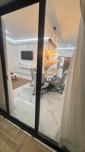 Een moderne woonkamer met een glazen schuindeur, een eethoek met een witte tafel en grijze stoelen, en een opvallende hanglamp.