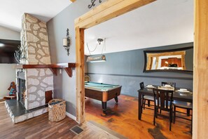Ein Billardtisch in einem Raum mit Kamin aus Stein, Holzbalken und einer Essecke mit Tisch und Stühlen, gedeckt für eine Mahlzeit.
