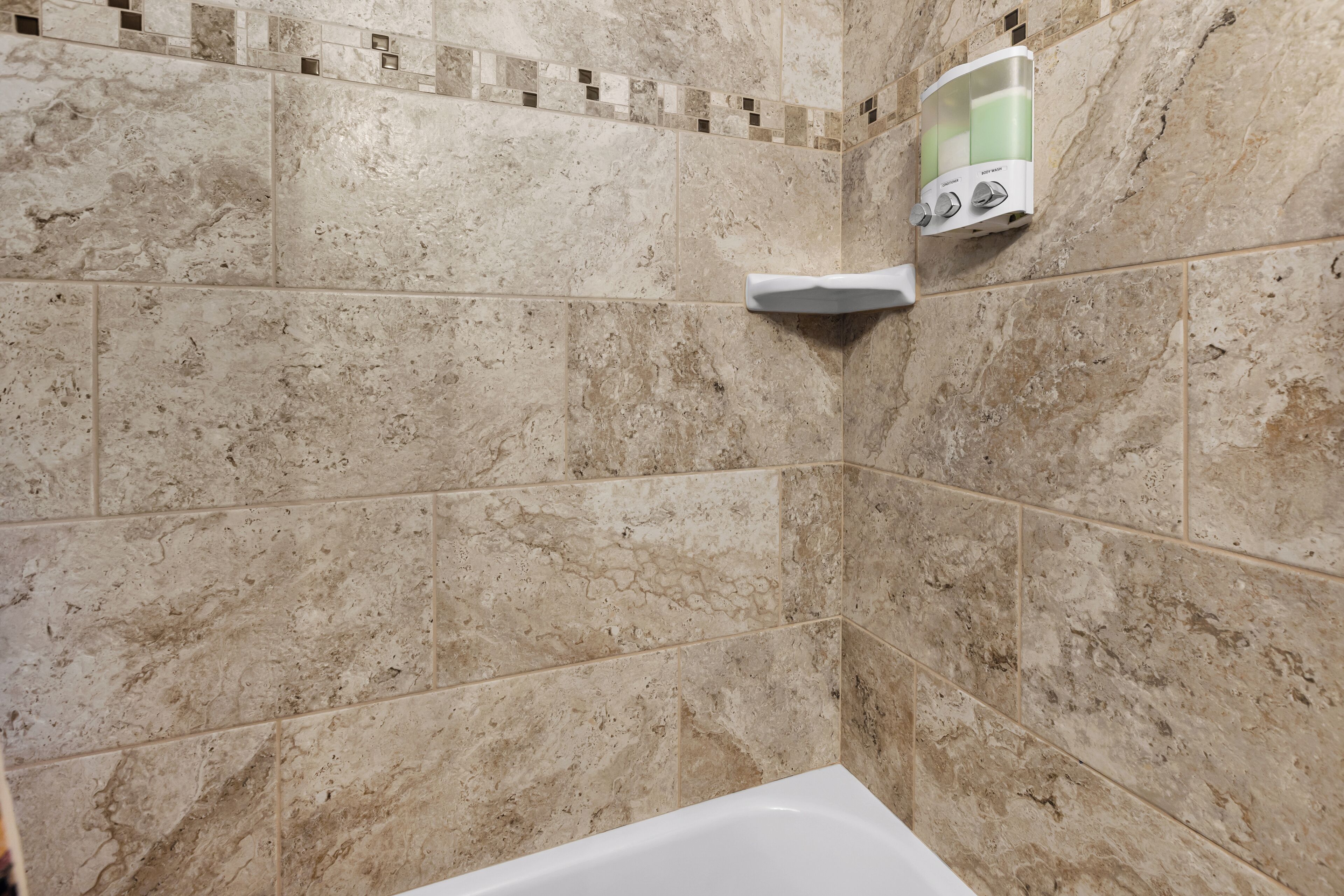 Ein Badezimmer mit beige farbenen Marmorfliesen, einer weißen Badewanne und einem Seifenspender an der Wand.