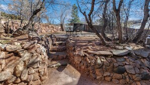 Um alpendre de pedra com uma mesa e cadeiras, rodeado por árvores e um muro de pedra.