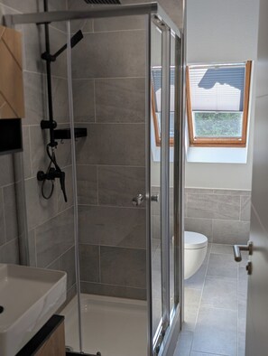 A modern bathroom with a glass-enclosed shower, a toilet, and a window with blinds. - Completely renovated design apartment close to the city (Lünen)