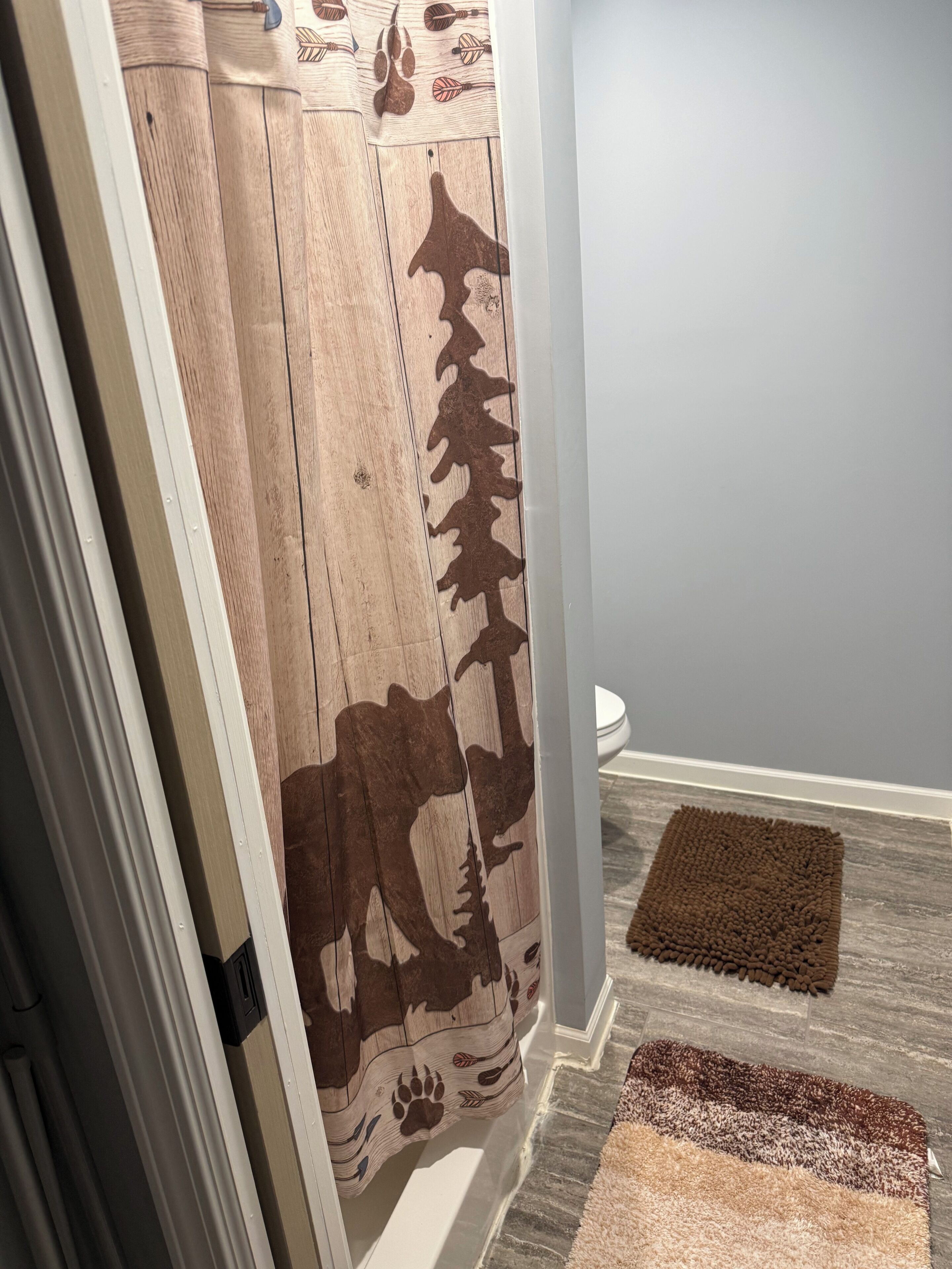 A bathroom with a wooden shower curtain, a toilet, and two rugs on the floor.