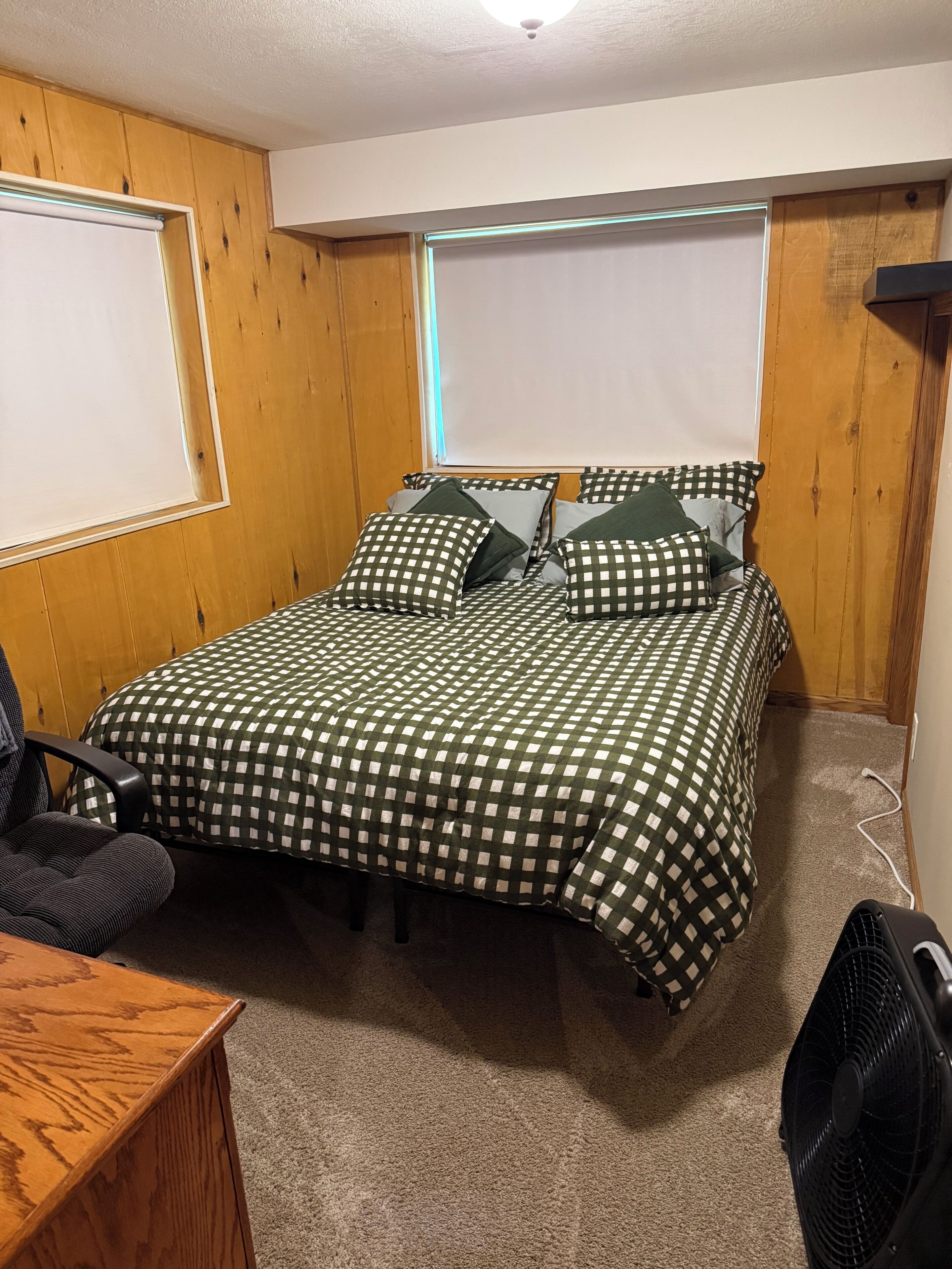 A bedroom with a checkered bedspread, a desk, and a fan.