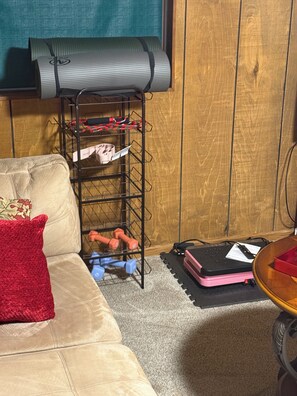 Una habitación con un estante metálico negro que contiene varios objetos, un sofá color beige con un cojín rojo y una pequeña mesa redonda con un objeto rosa encima.