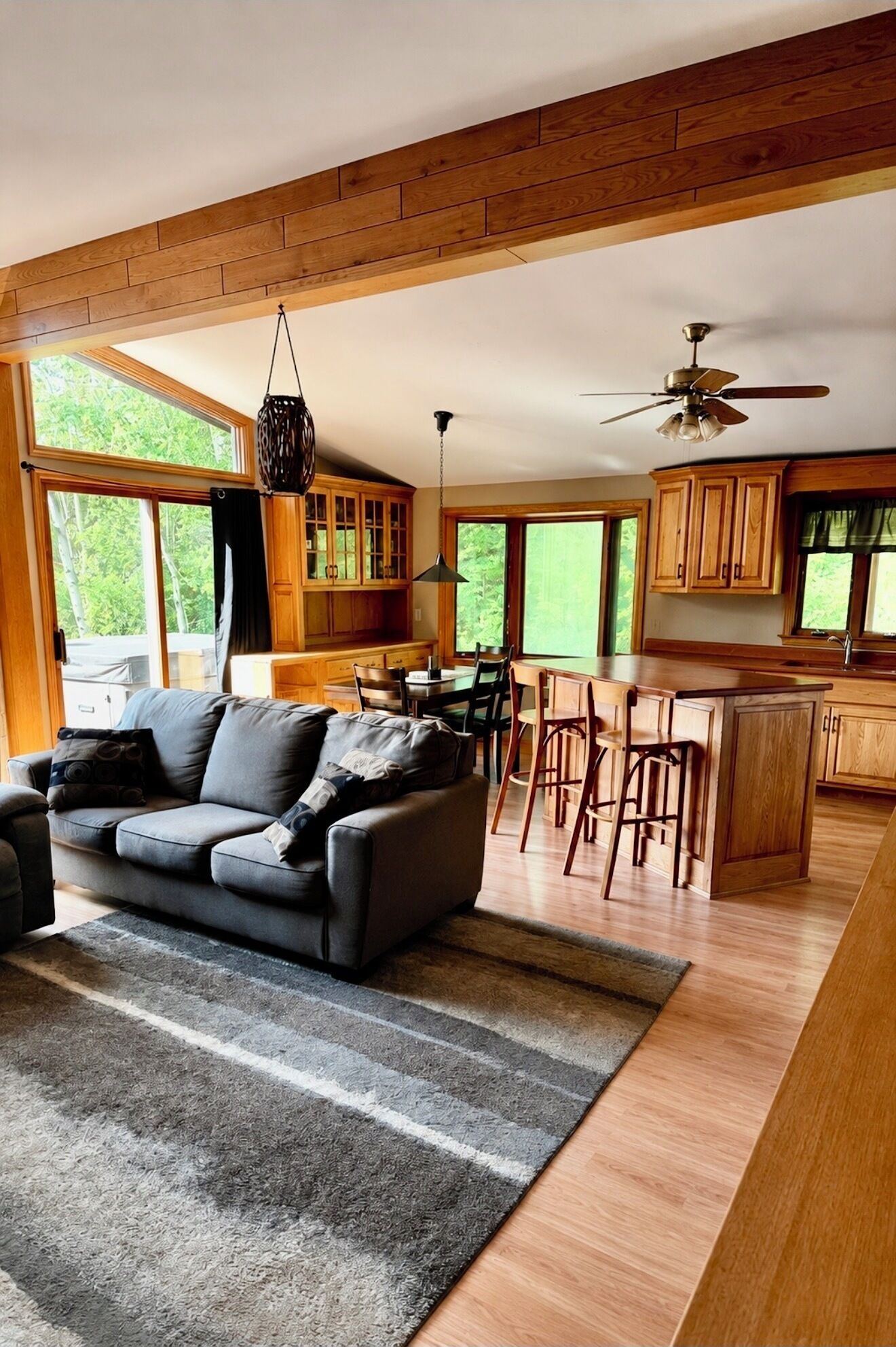 A modern living room with a sofa, a dining area with chairs, a kitchen with wooden cabinets, and large windows offering a view of greenery.