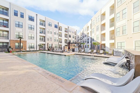 A swimming pool with lounge chairs and a waterfall feature, surrounded by multi-story buildings with balconies. - Luxurious 2B2B Near Toyota HQ (Frisco)