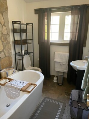 Une salle de bain avec une baignoire, des toilettes et un lavabo. Il y a une fenêtre avec des rideaux, un porte-serviettes et une étagère.