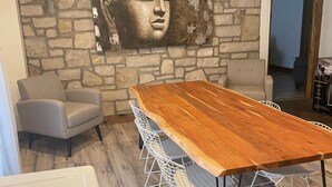 A modern dining area with a wooden table and white wireframe chairs, a stone wall with a Buddha painting, and a grey armchair. - The Quinta at Balm Beach (Perkinsfield)