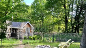 A cabin surrounded by a dense forest, with a fire pit and two Adirondack chairs in the foreground. - The Quinta at Balm Beach (Perkinsfield)